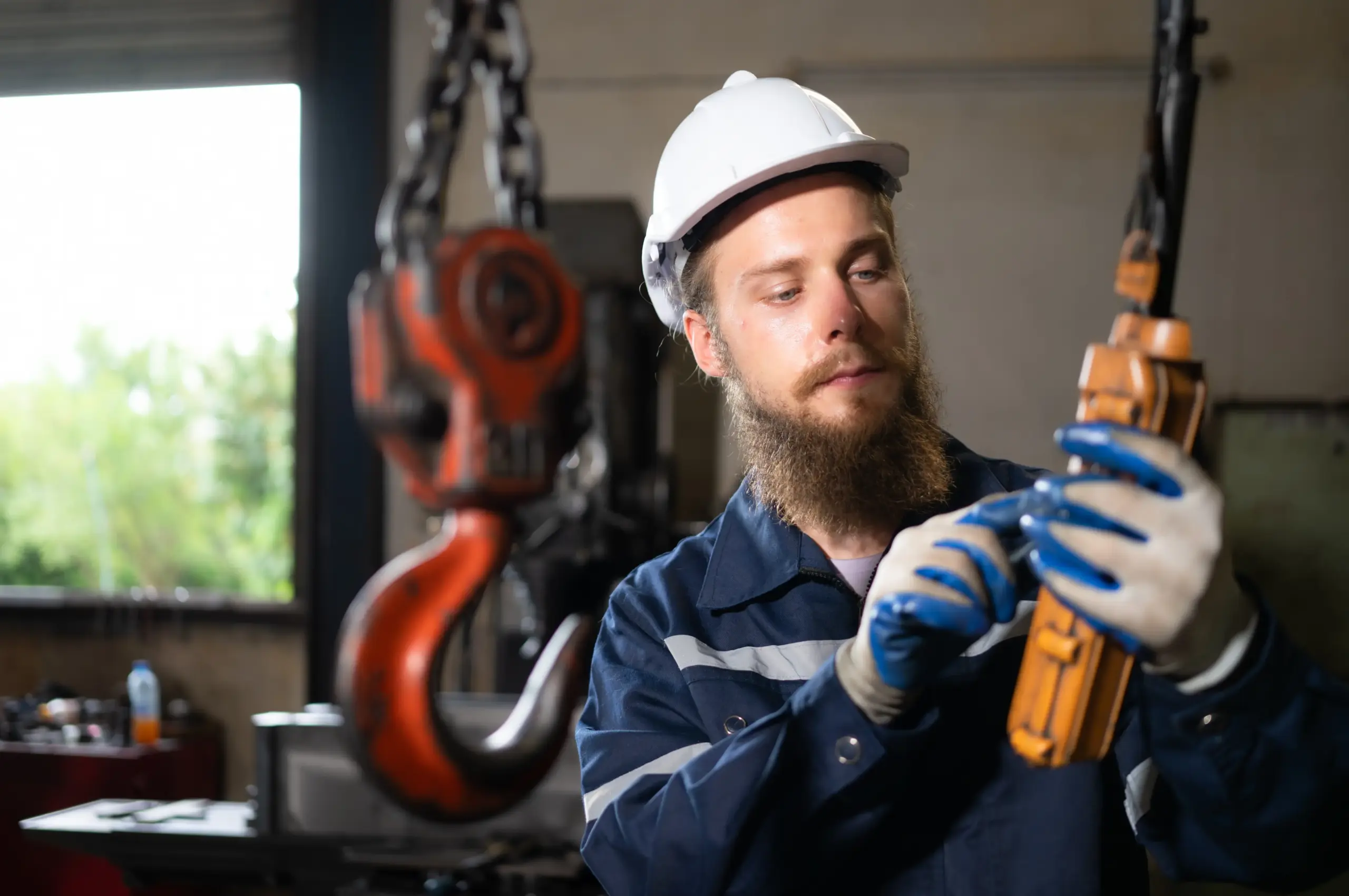 A bearded worker in safety gear and gloves operates a crane control panel beside a large red industrial hook inside a workshop or factory setting.