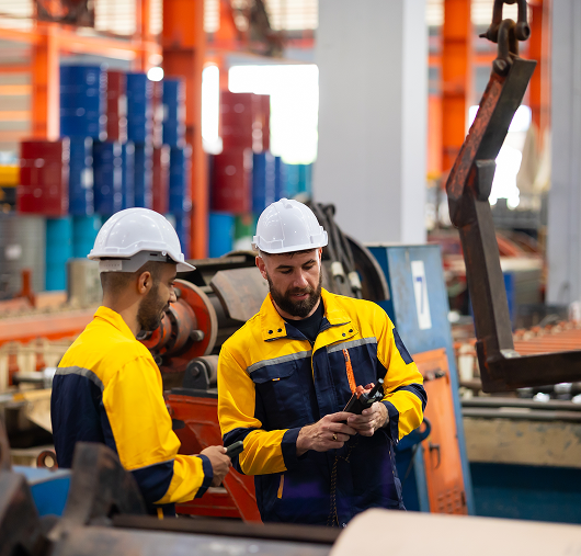 Two factory workers wearing yellow and navy uniforms and white helmets stand in an industrial setting, examining equipment and discussing something while surrounded by machinery and colorful storage bins.