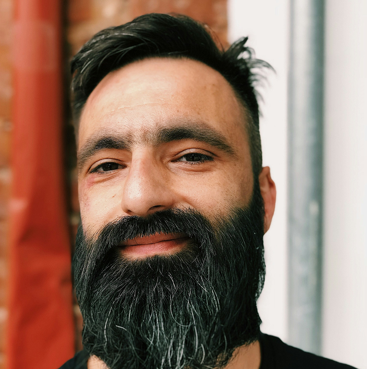 Close-up of a man with a thick, dark beard and mustache, short dark hair, and a slight smile. The background includes blurred red, brick, and white elements.