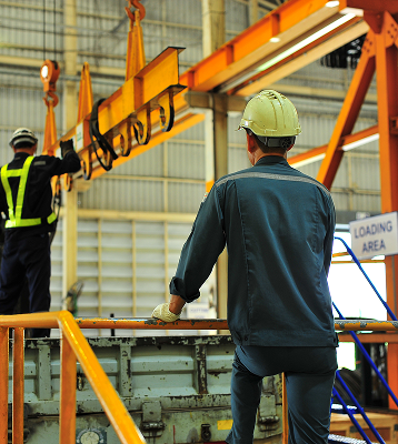 Two workers in safety gear operate a large overhead crane inside an industrial facility. One stands on a platform watching, while the other operates controls near a Loading Area sign.