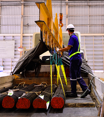 A worker in safety gear operates a crane to lift bundled steel rods onto a truck inside an industrial warehouse.