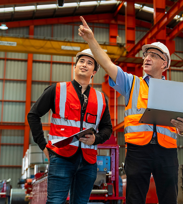Two workers in safety vests and hard hats stand in a factory; one holds a laptop and points upward while the other holds a clipboard, both looking in the direction indicated.