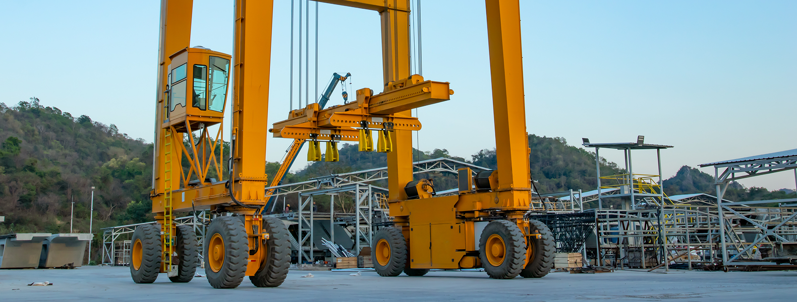 A large yellow gantry crane stands on a concrete surface at an industrial site, surrounded by steel structures and hills in the background under a clear sky.