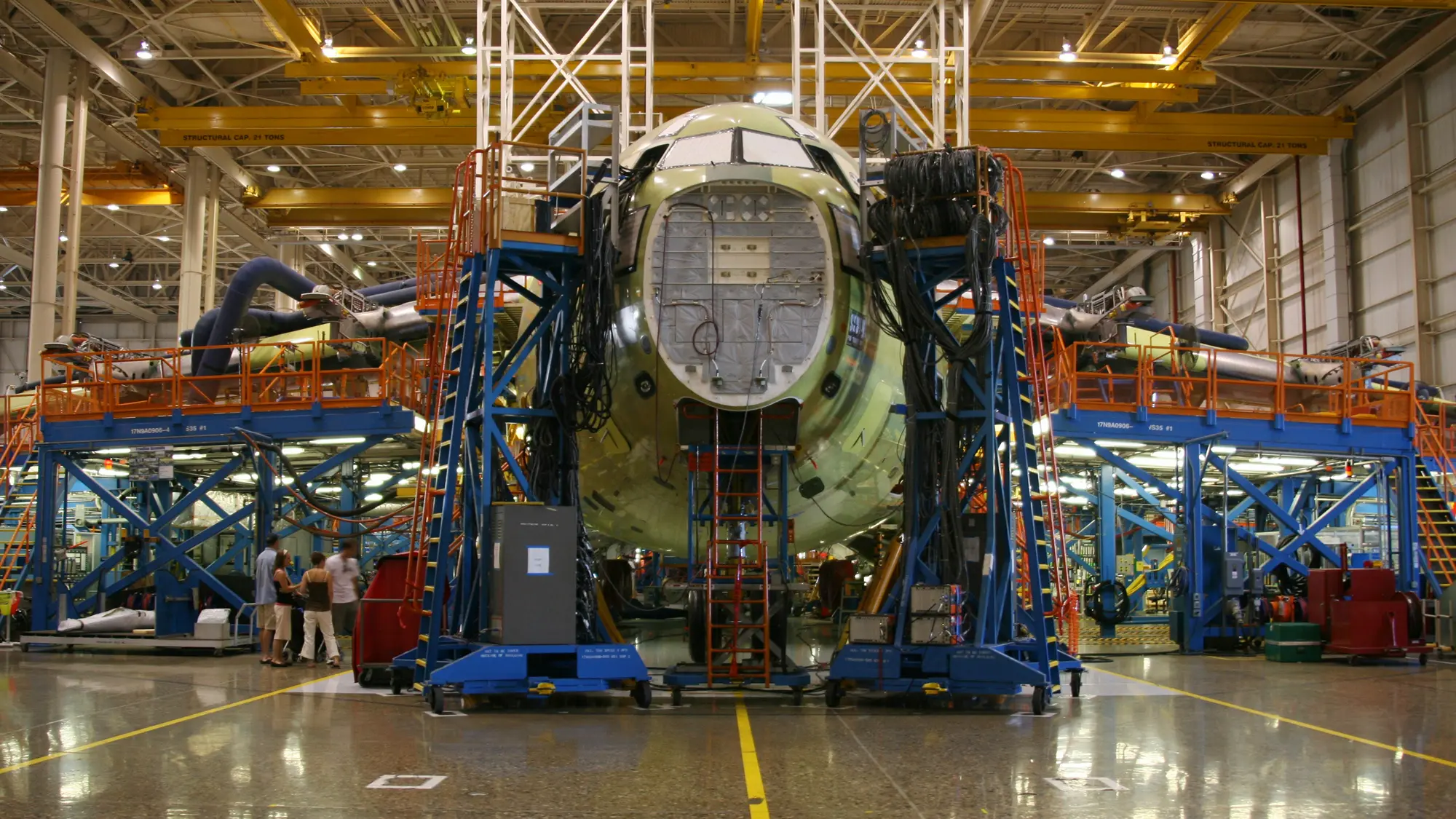 An airplane under construction inside a large factory, surrounded by scaffolding, machinery, and workers. The nose of the plane is open, revealing internal components and cables. Bright lights illuminate the spacious industrial setting.