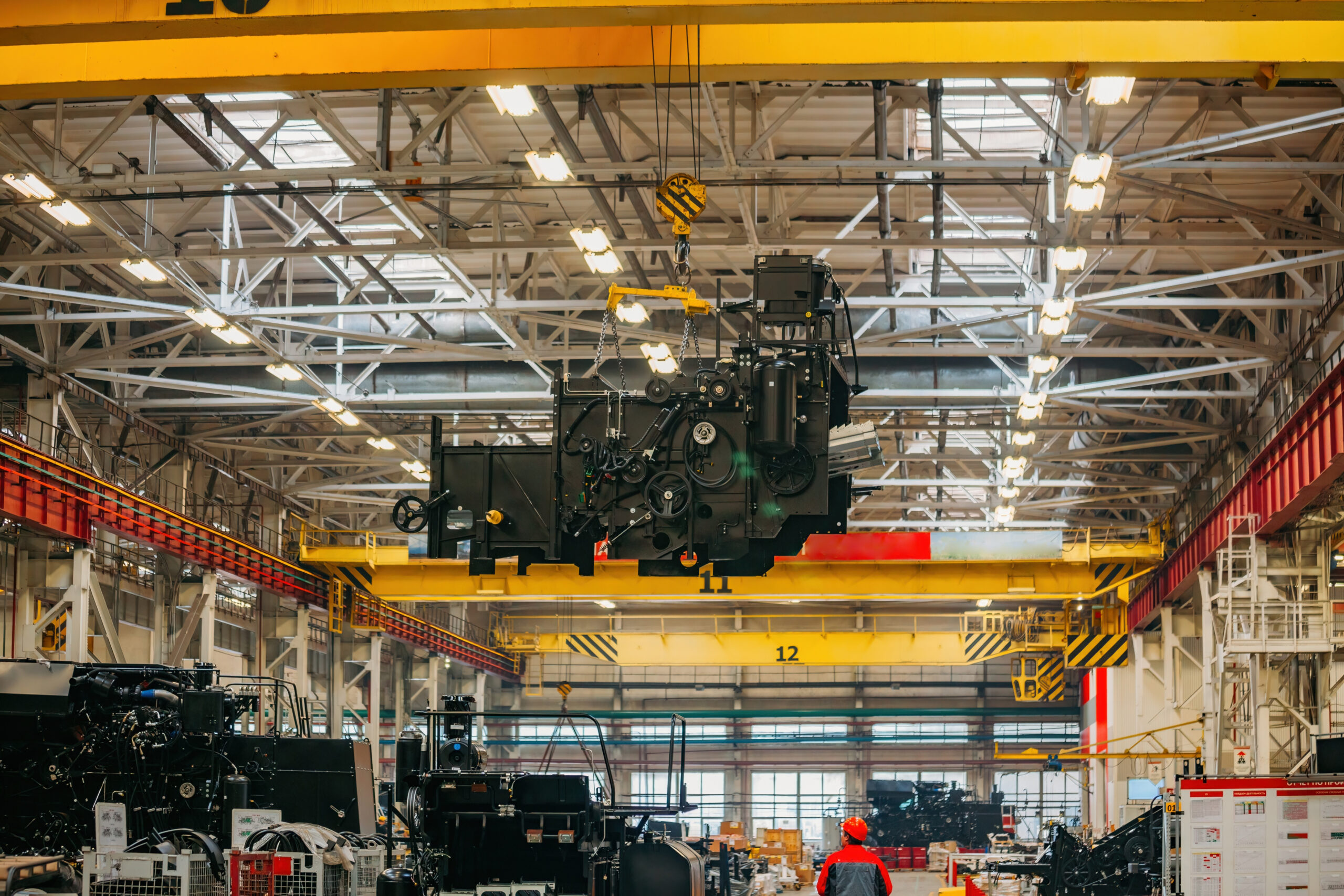 A large industrial machine is being lifted by an overhead crane inside a spacious, brightly lit factory. A worker in a red helmet stands below, observing the process.