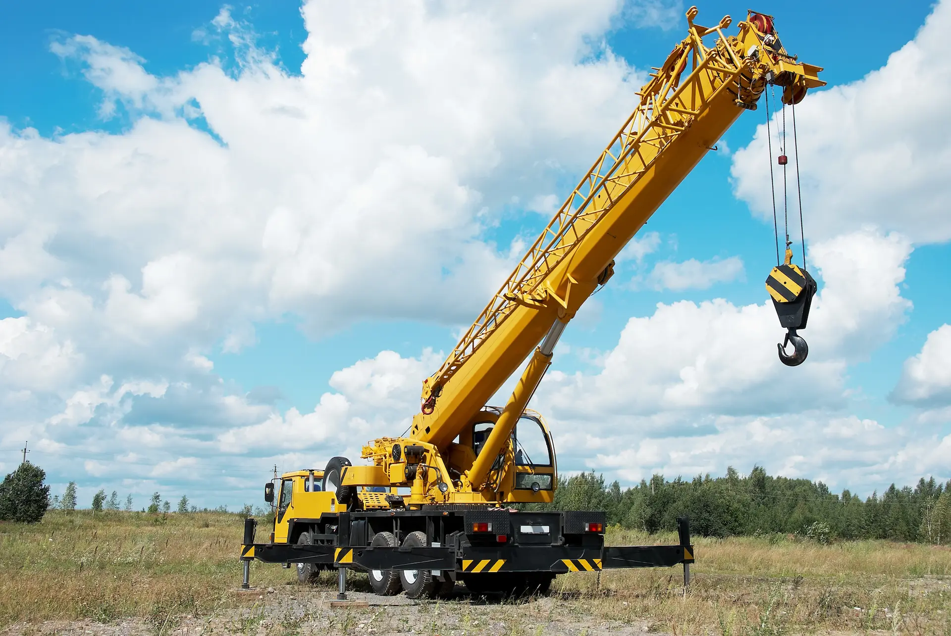 A large yellow mobile crane with an extended boom is parked on grassy terrain under a blue sky with scattered clouds. The crane’s stabilizers are deployed, and the hook is suspended in the air. Trees are visible in the background.