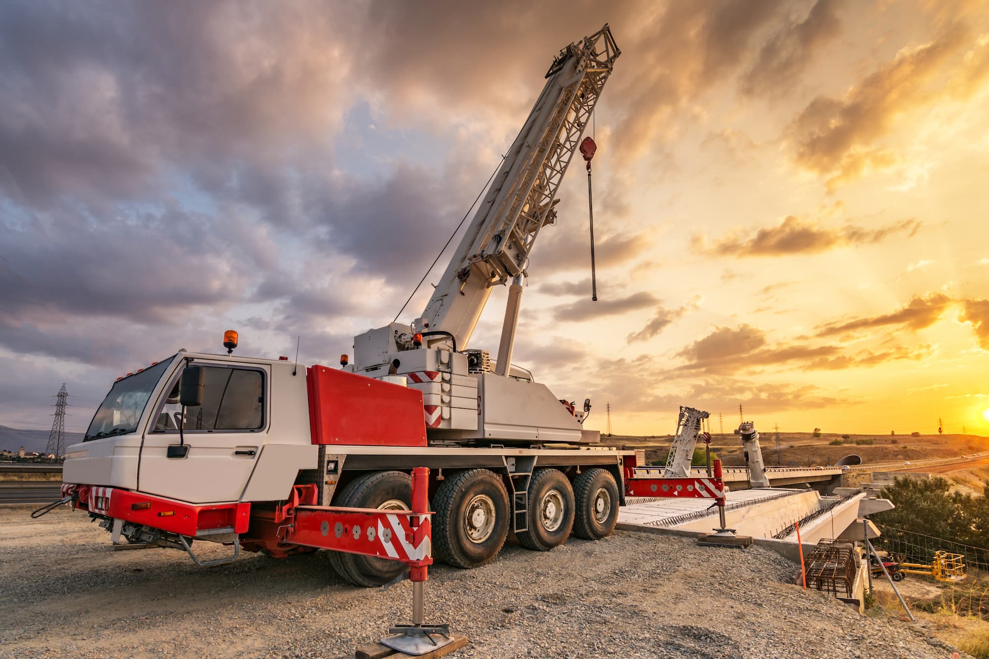 A large mobile crane with an extended boom is parked on a construction site at sunset, with a dramatic cloudy sky and distant hills in the background.