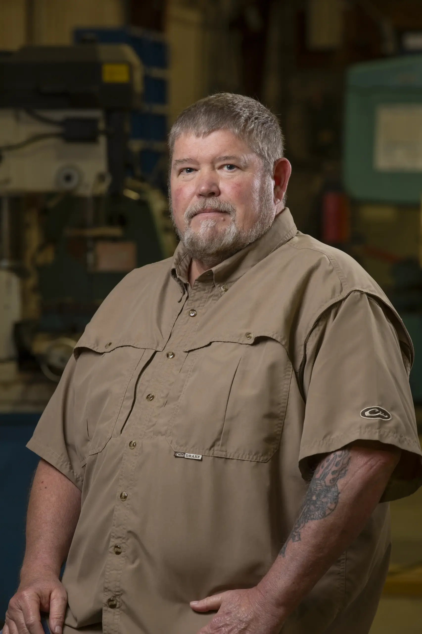 A middle-aged man with short gray hair and a beard, wearing a tan button-up shirt, stands indoors with industrial equipment in the blurred background. He has a tattoo on his right forearm and looks calmly at the camera.