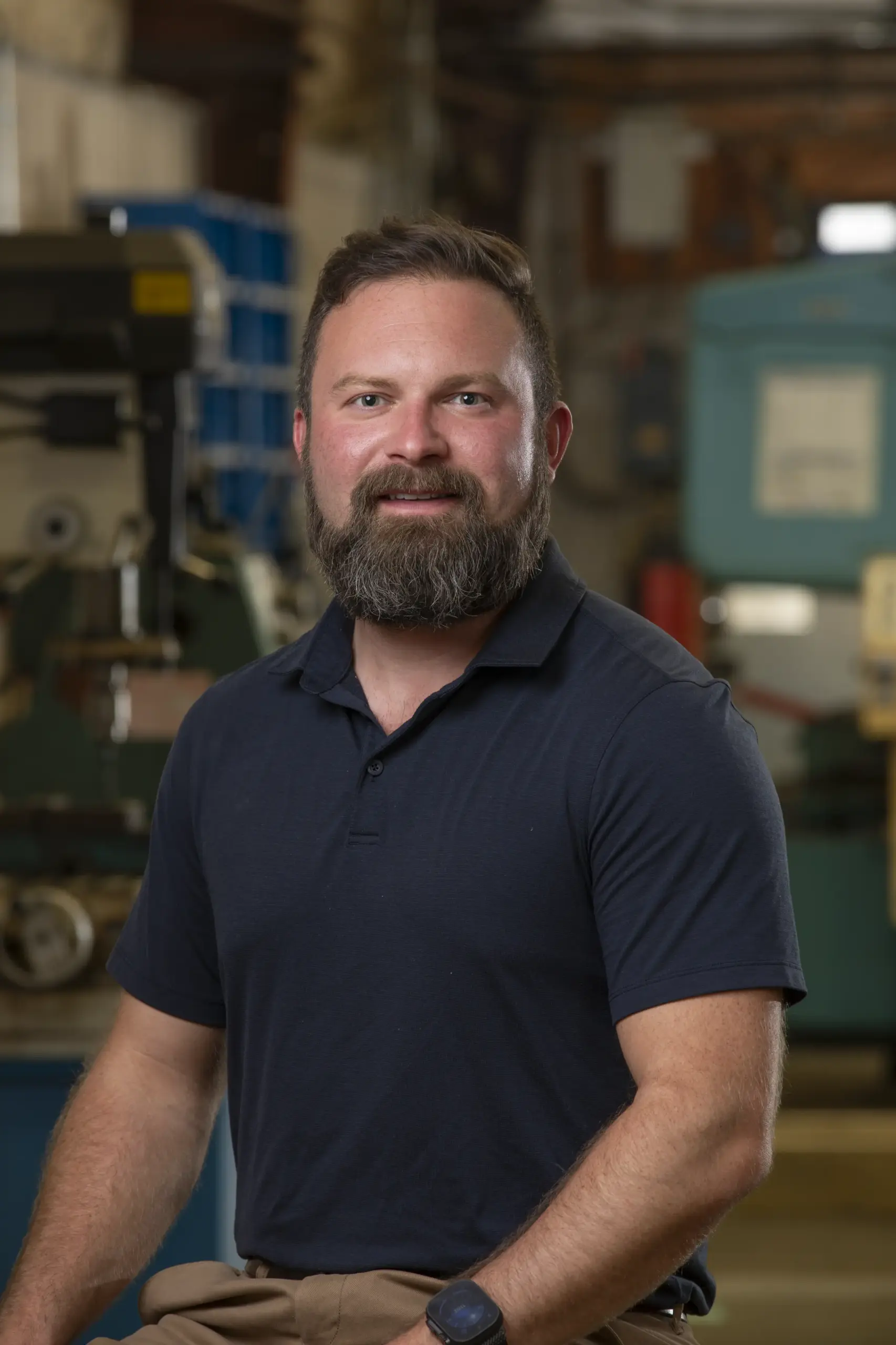 A bearded man in a navy blue polo shirt sits and smiles in an industrial workshop setting, with machinery and equipment visible in the background.