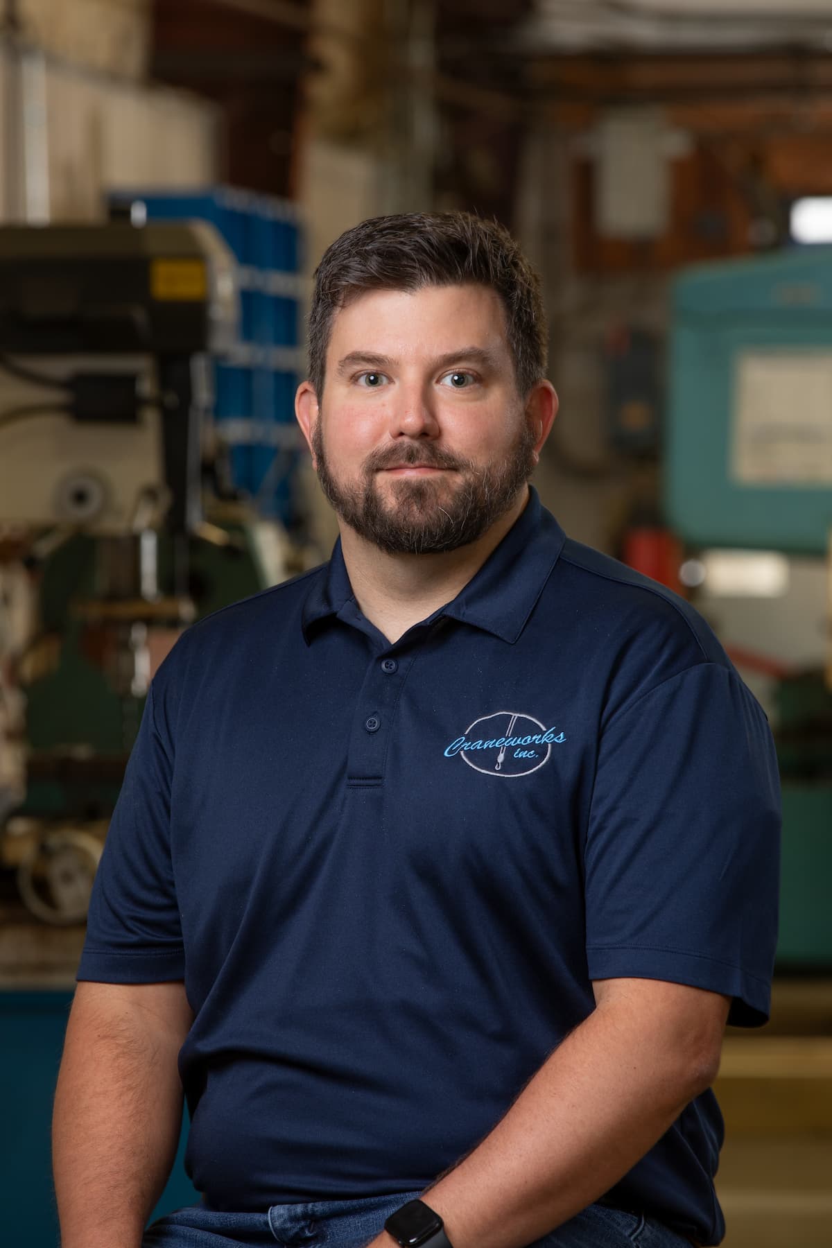 A man with short dark hair and a beard, wearing a navy blue polo shirt with a logo, sits and faces the camera in an industrial workshop setting with machines in the background.