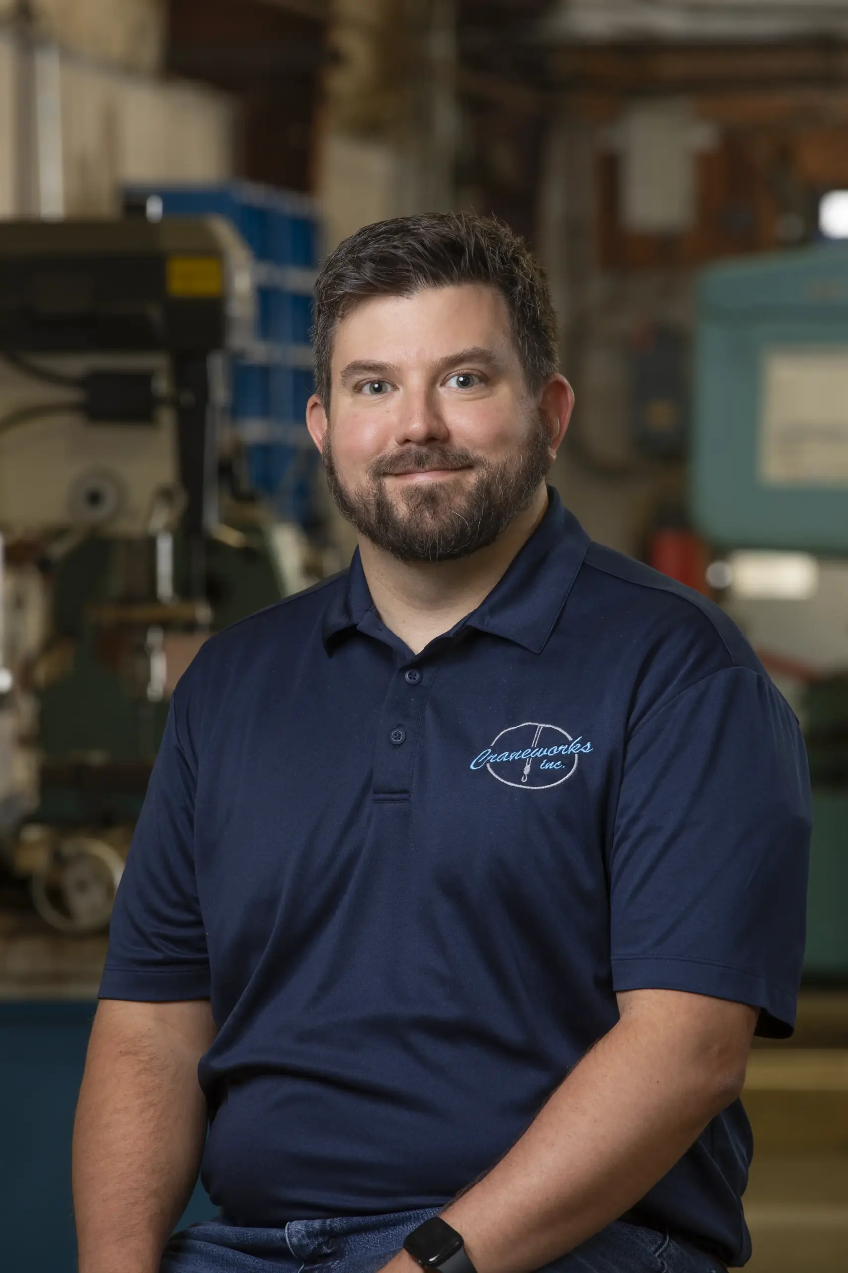 A man with short brown hair and a beard, wearing a navy blue polo shirt with a logo, sits and smiles in an industrial workshop setting with blurred machinery in the background.