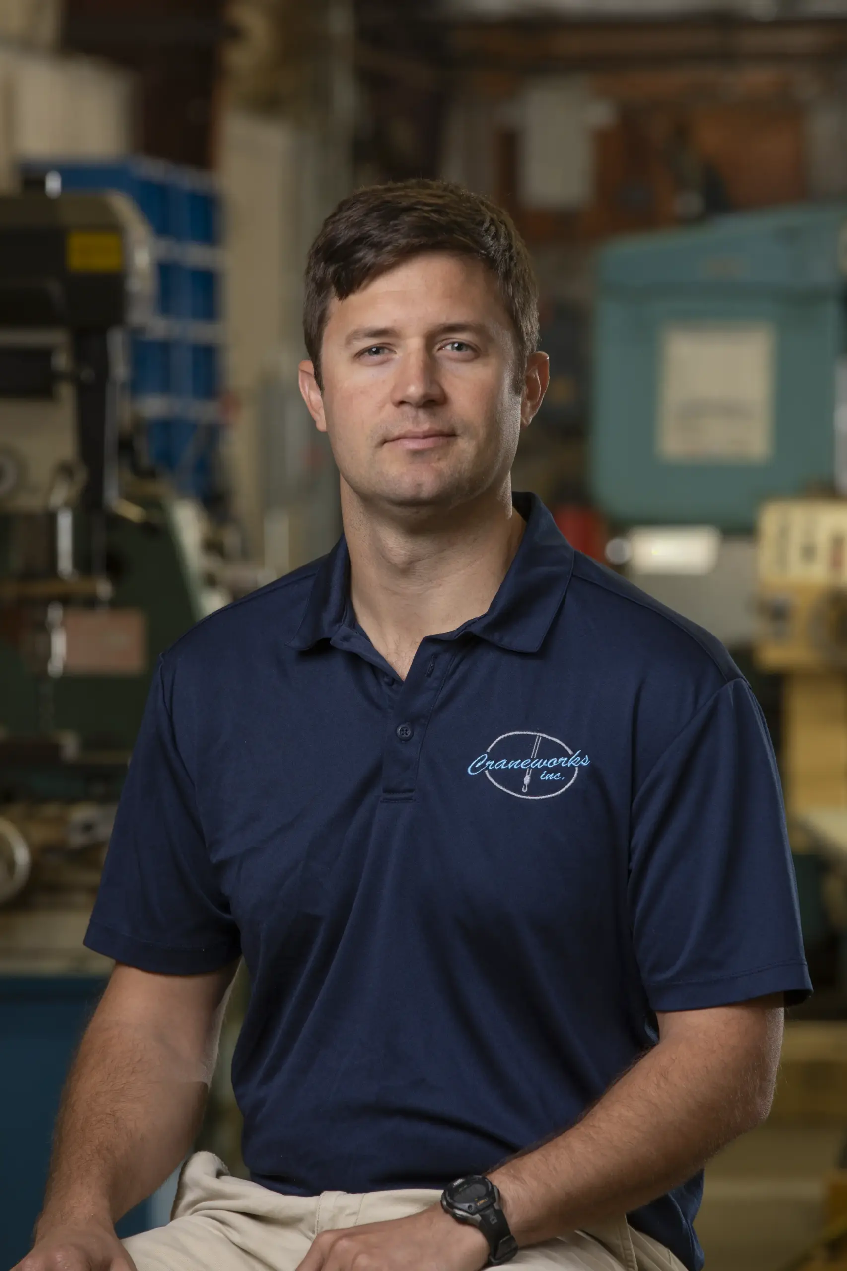 A man with short brown hair wearing a navy blue polo shirt and khaki pants sits indoors in an industrial or workshop setting, looking at the camera with a neutral expression.