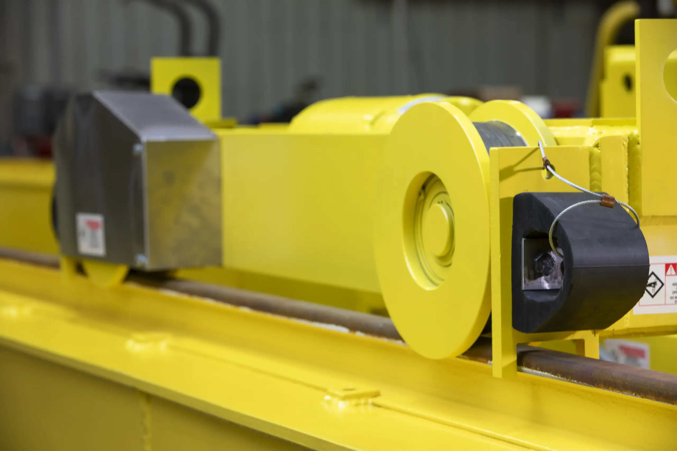 Close-up of a bright yellow industrial overhead crane mechanism with a metal cable wound around a spool and various mechanical components in a warehouse setting.