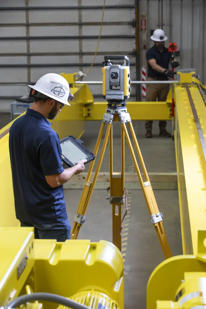 Two workers in hard hats use surveying equipment and a tablet inside an industrial facility with yellow machinery and metal structures. One operates a tripod-mounted device while the other stands in the background.