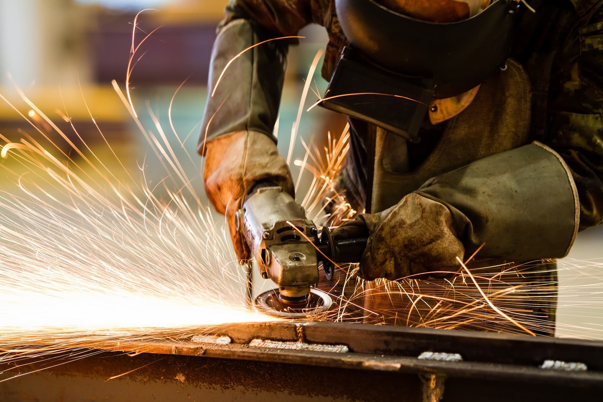 A person wearing protective gloves and a welding mask uses an angle grinder on a metal surface, producing bright, flying sparks in a workshop setting.