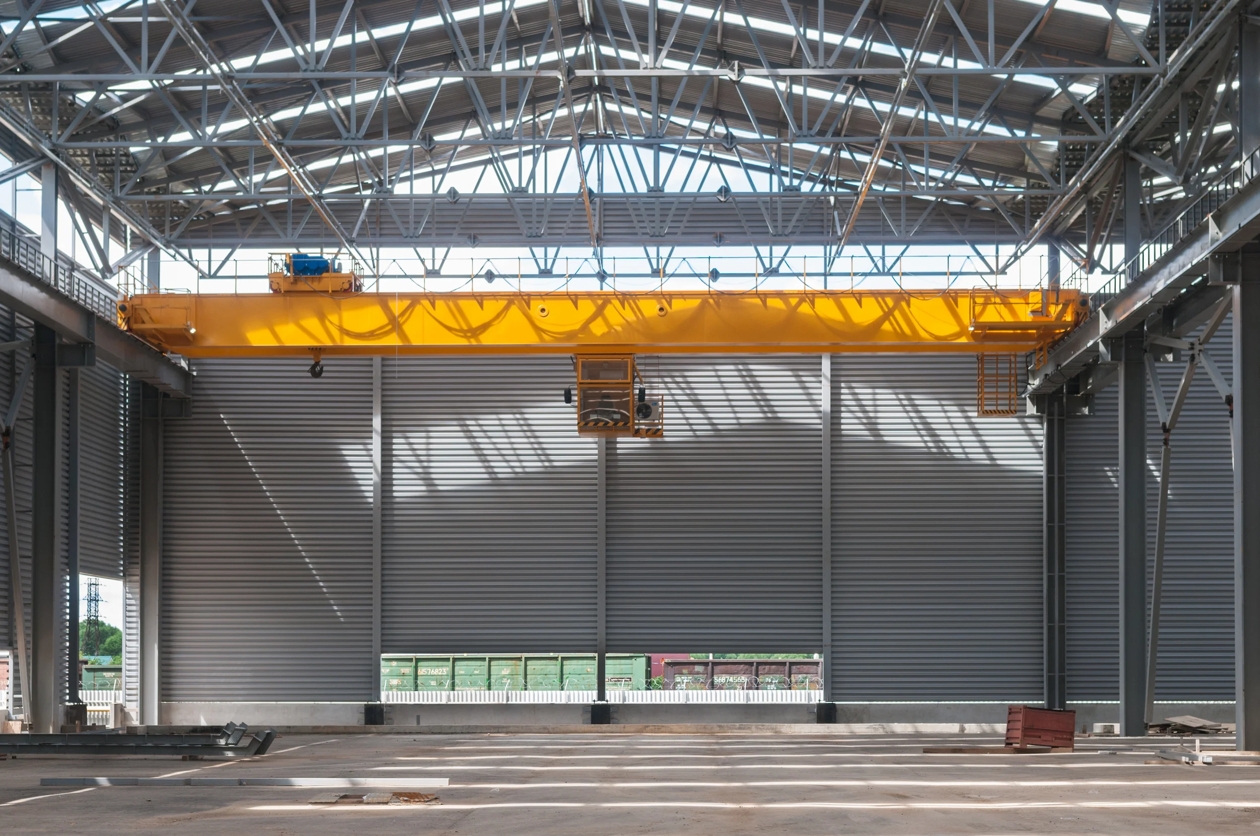 Interior of a large industrial warehouse with a high metal roof and steel beams, featuring a bright yellow overhead crane. The space is empty, with a few items on the concrete floor and sunlight streaming in.
