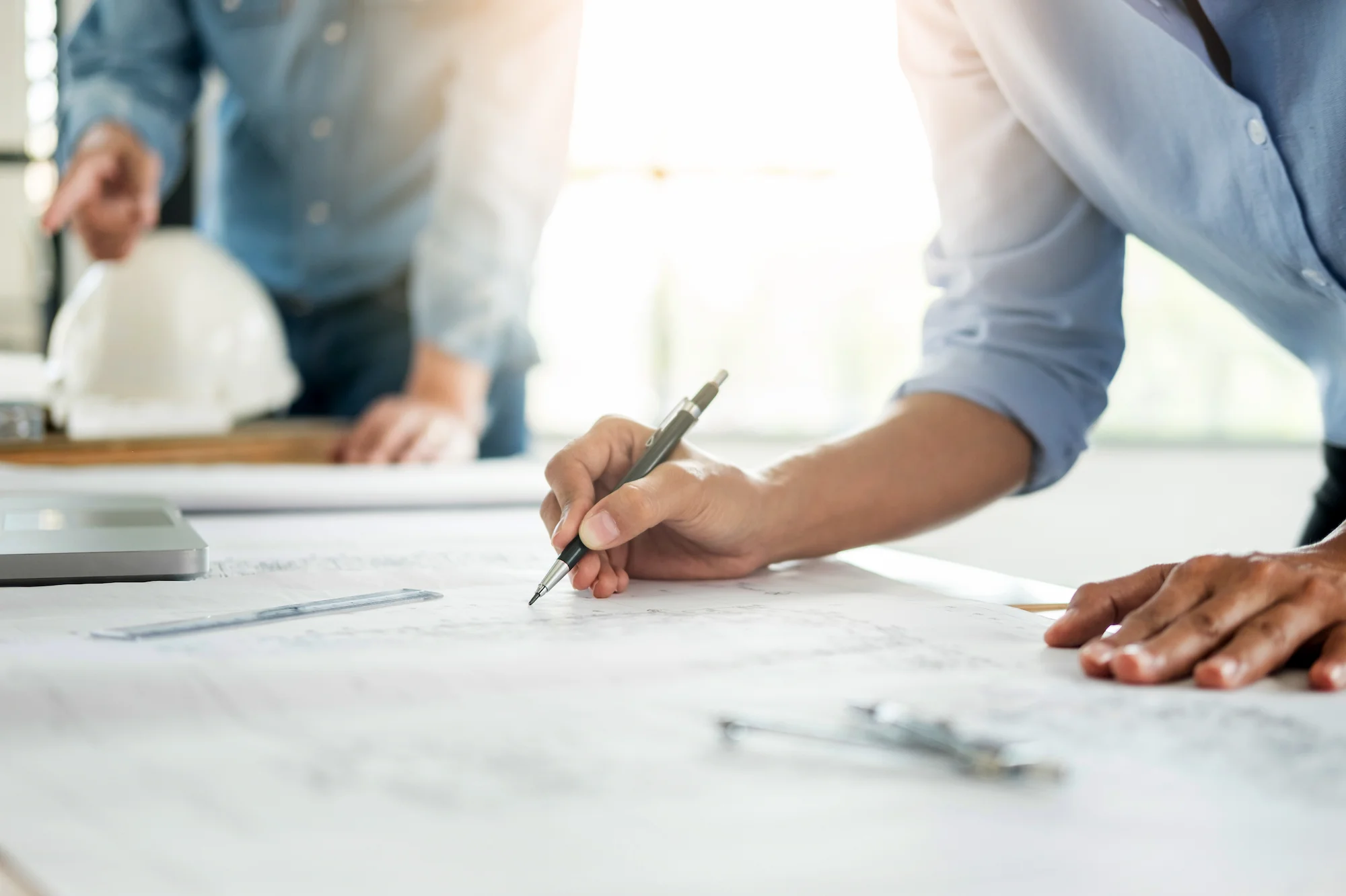 Close-up of a person’s hand drawing or writing on architectural blueprints on a desk, with another person and a white safety helmet blurred in the background. The scene is well-lit with natural light.