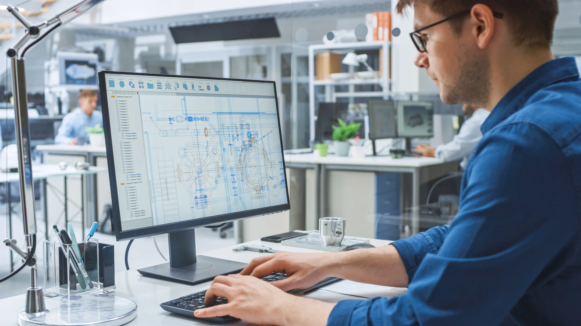 A man wearing glasses and a blue shirt sits at a desk working on a computer with engineering blueprints displayed on the monitor in a modern office with other people in the background.