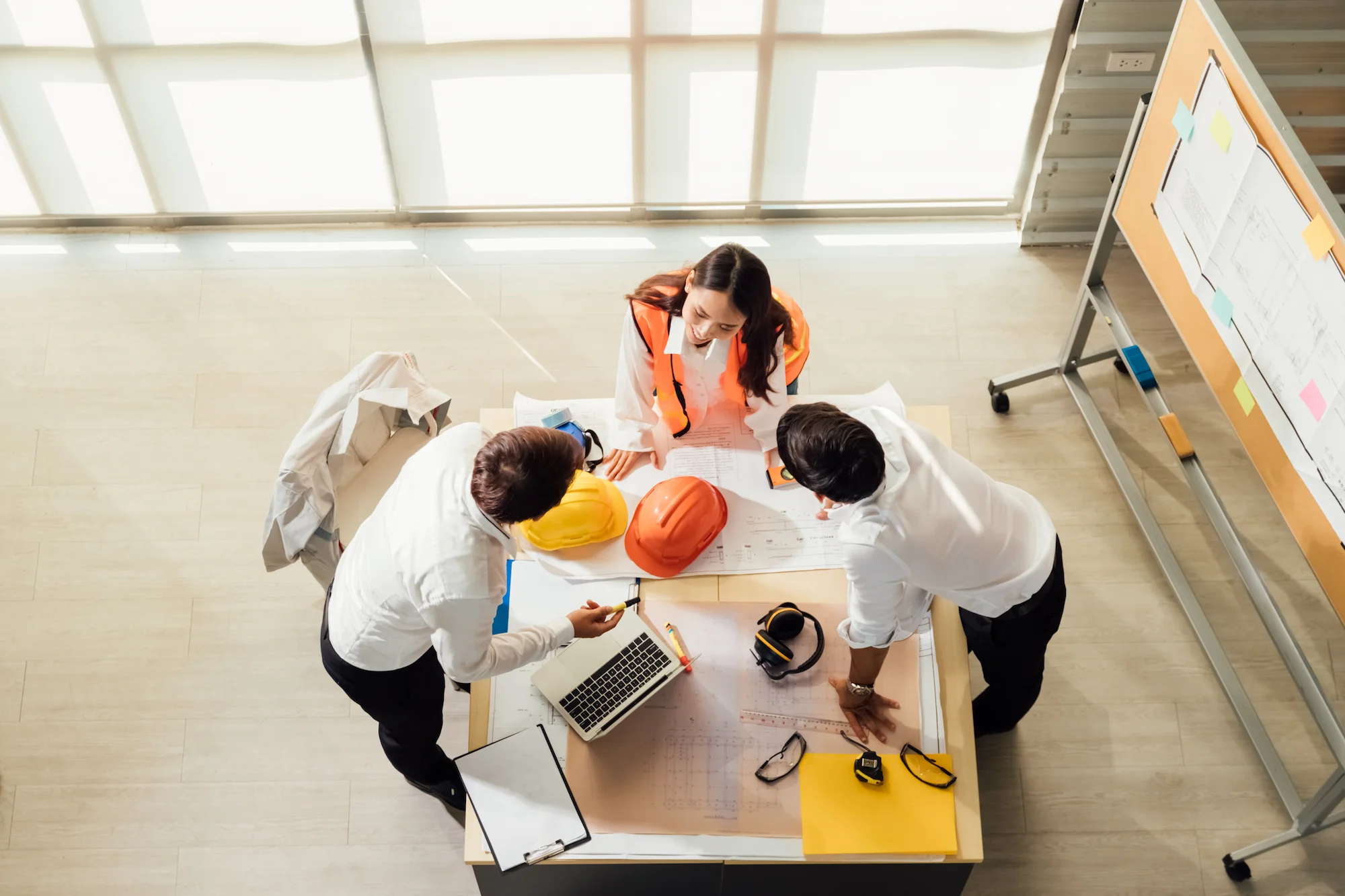 Three people, two in white shirts and one in an orange safety vest, stand around a table with blueprints, hard hats, a laptop, and safety glasses, collaborating on a construction project.