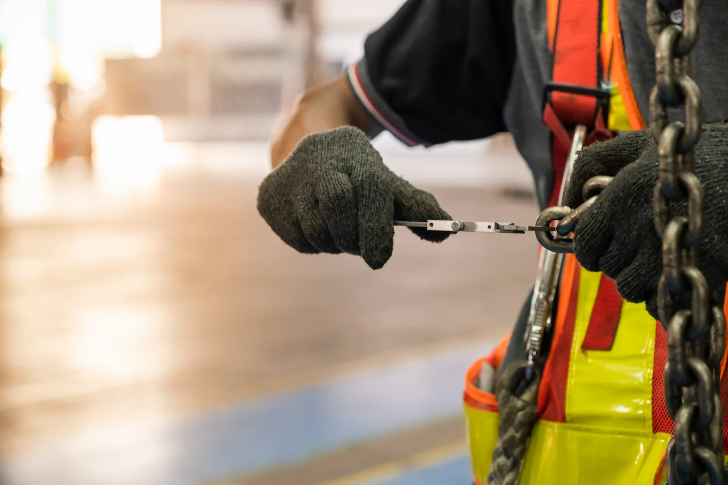 A person wearing black gloves and a safety vest uses a tool to secure a metal hook or carabiner to a chain, likely performing maintenance or safety checks in an industrial setting.