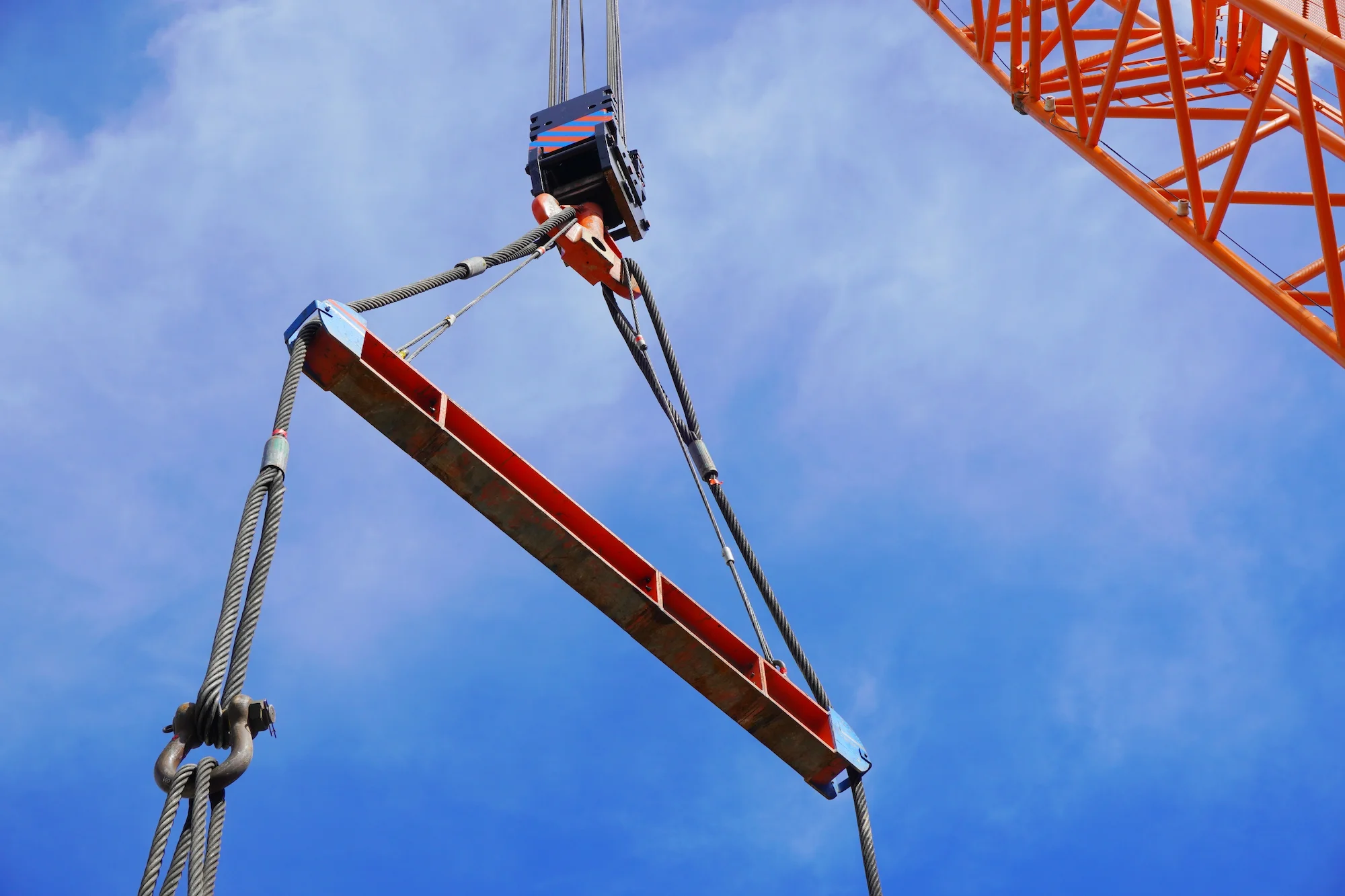 A close-up view of a crane lifting a long red steel beam with cables against a bright blue sky, showing part of the cranes orange structure and pulley system.