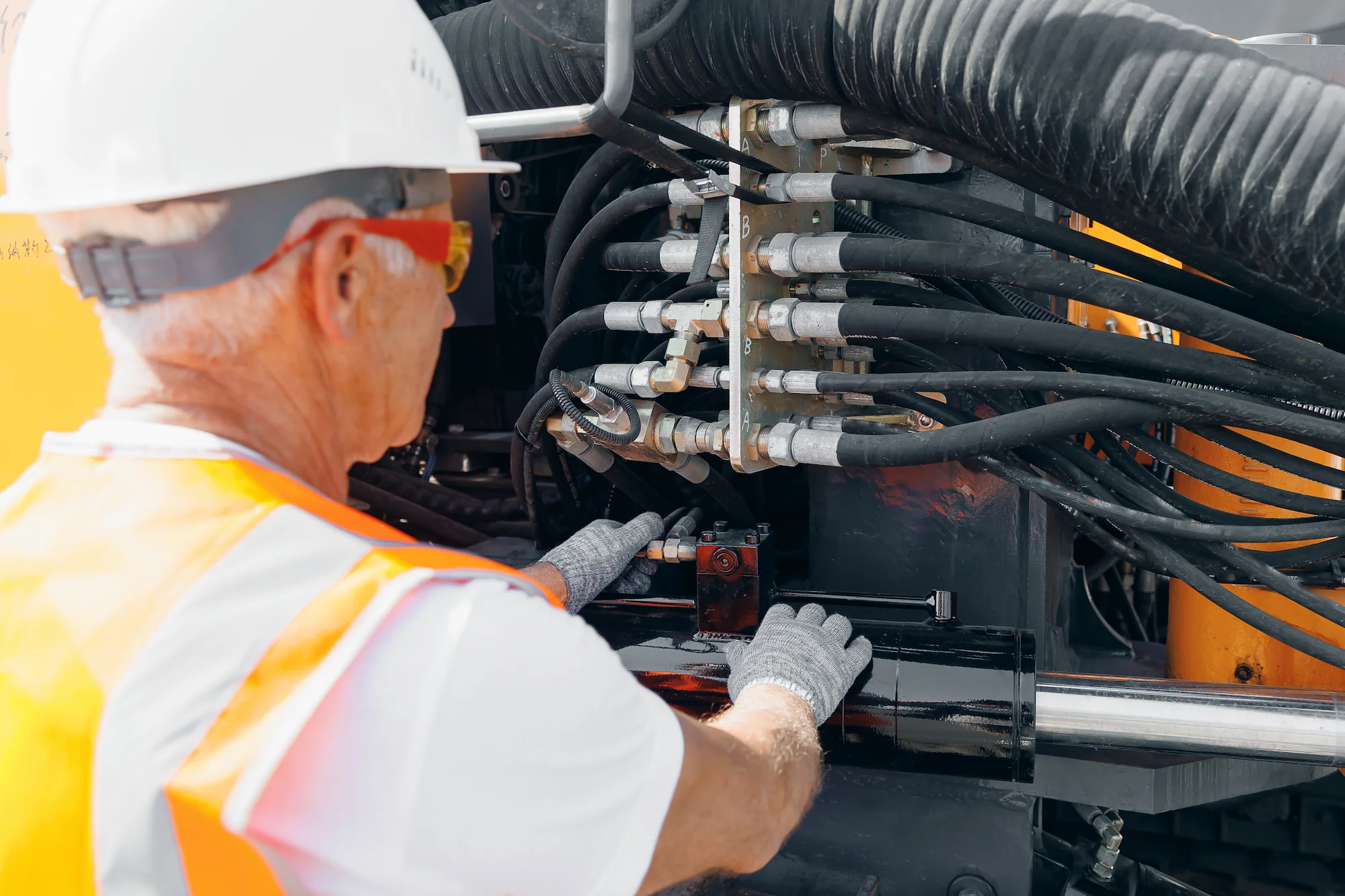 A construction worker wearing a white hard hat, safety glasses, gloves, and an orange vest operates machinery by adjusting controls and hoses on a piece of heavy equipment.