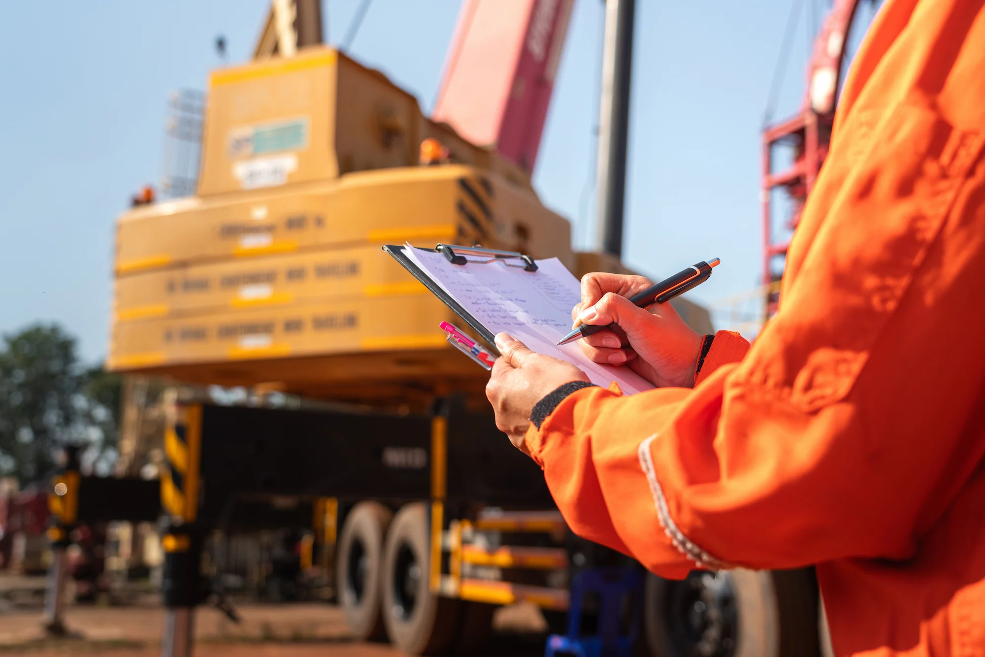 A person in an orange safety uniform writes on a clipboard at a construction site, with heavy machinery and cranes visible in the background.
