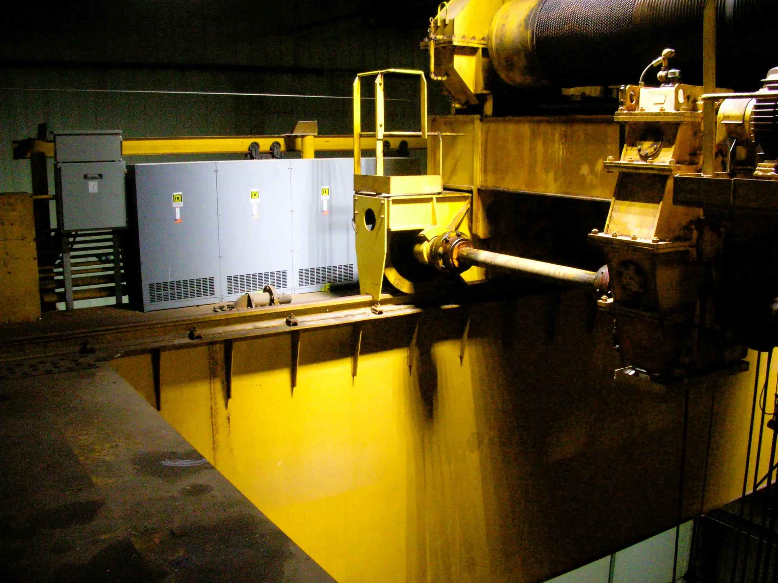 Large yellow industrial crane machinery indoors, with control panels and thick metal beams visible. The area is lit by artificial light, highlighting mechanical details and metal surfaces.