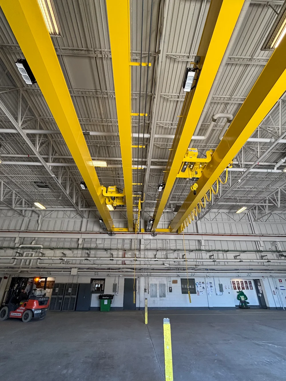 Interior of an industrial warehouse with yellow overhead cranes attached to the ceiling. The spacious area has concrete floors, machinery, and vehicles visible along the walls. Bright lighting illuminates the space.
