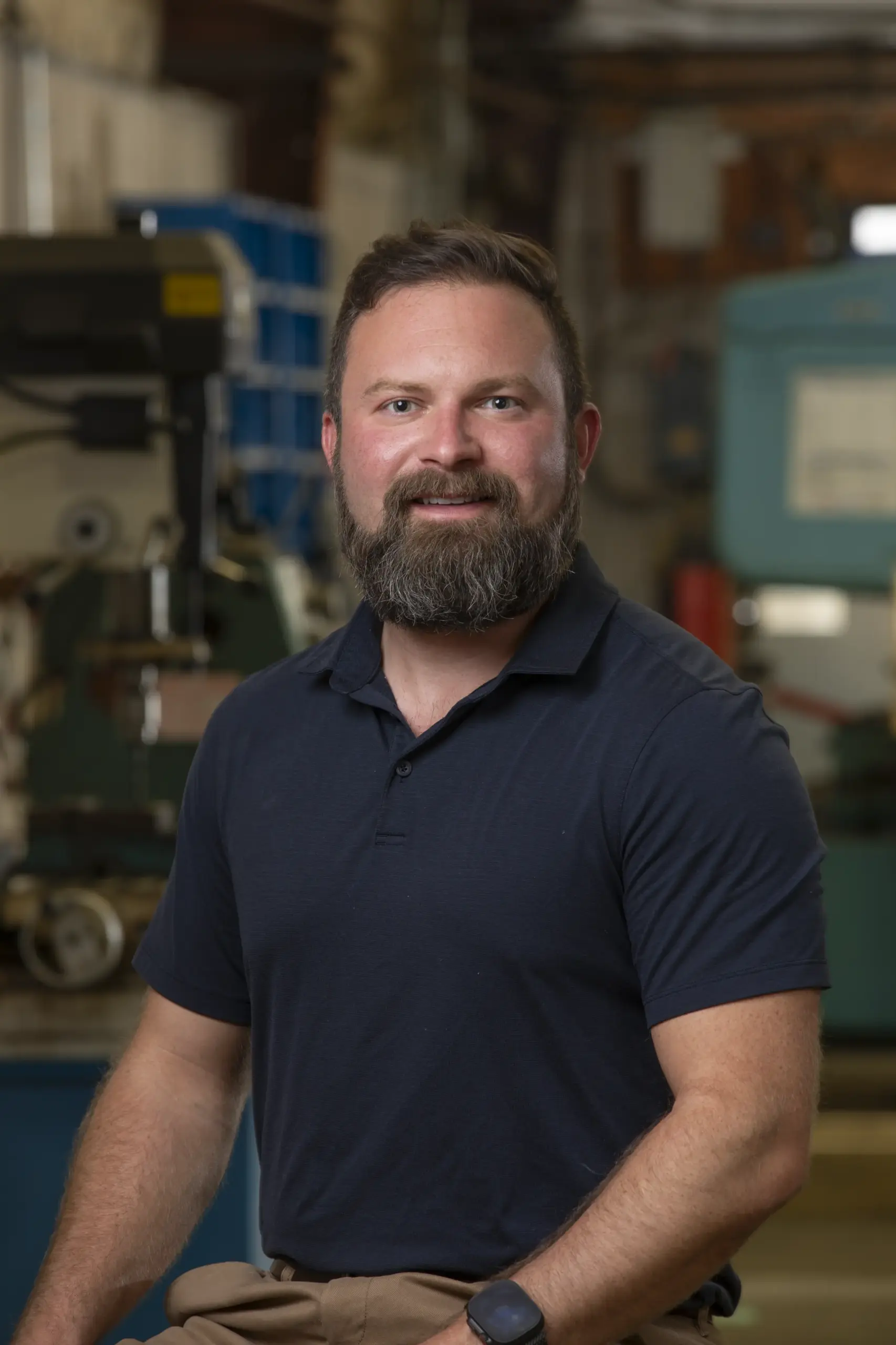 A bearded man wearing a dark polo shirt and watch poses and smiles in an industrial workshop setting, with machinery and tools visible in the background.