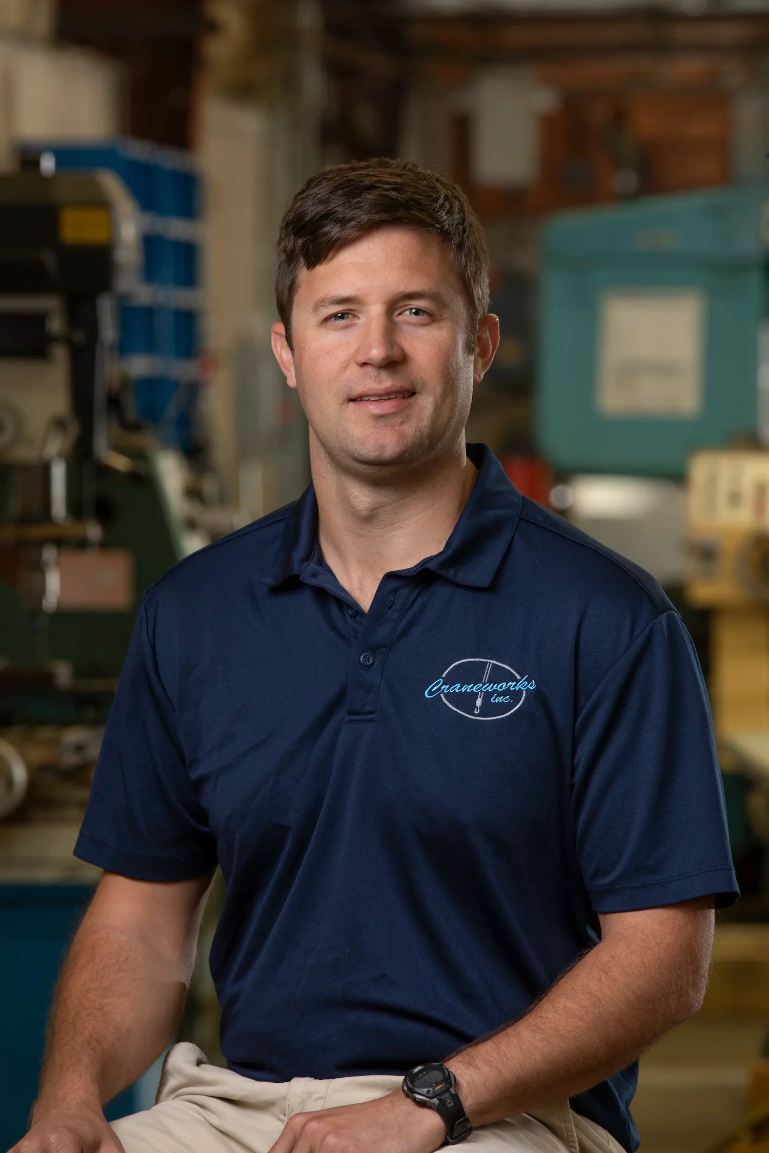 A man with short brown hair wearing a navy blue polo shirt sits and smiles in an industrial or workshop setting, with machinery and equipment visible in the background.
