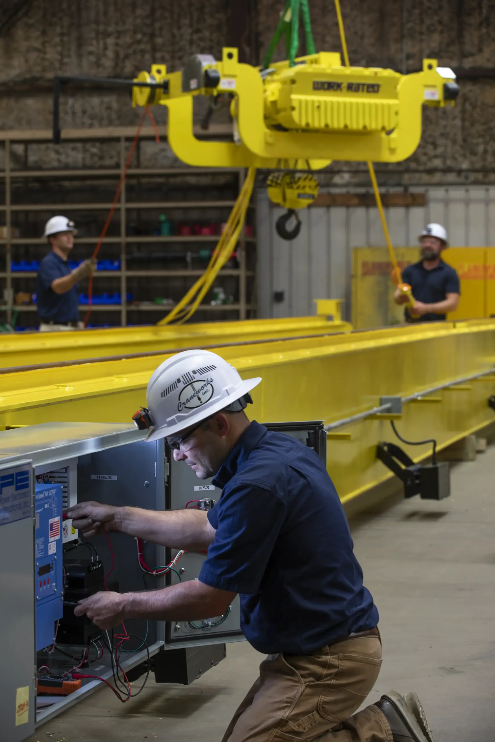 A worker in a hard hat adjusts wiring inside a control panel near a large yellow industrial crane, while two other workers operate the crane in the background inside a warehouse.