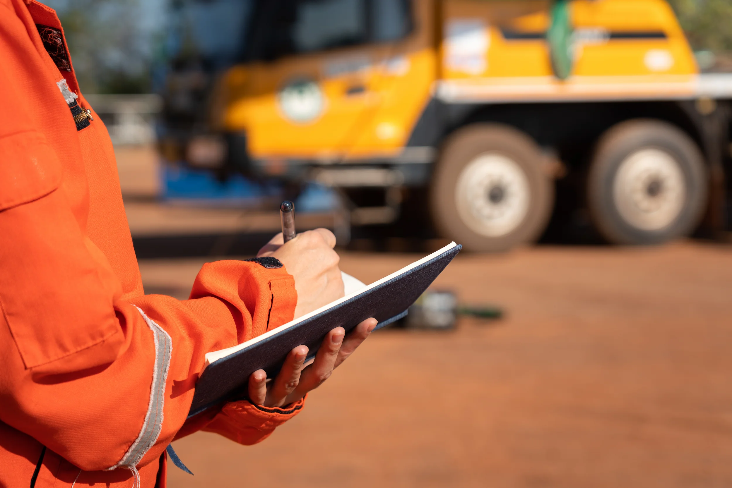 Person in an orange safety uniform writes in a notebook outdoors, with a blurred yellow construction truck and equipment in the background.