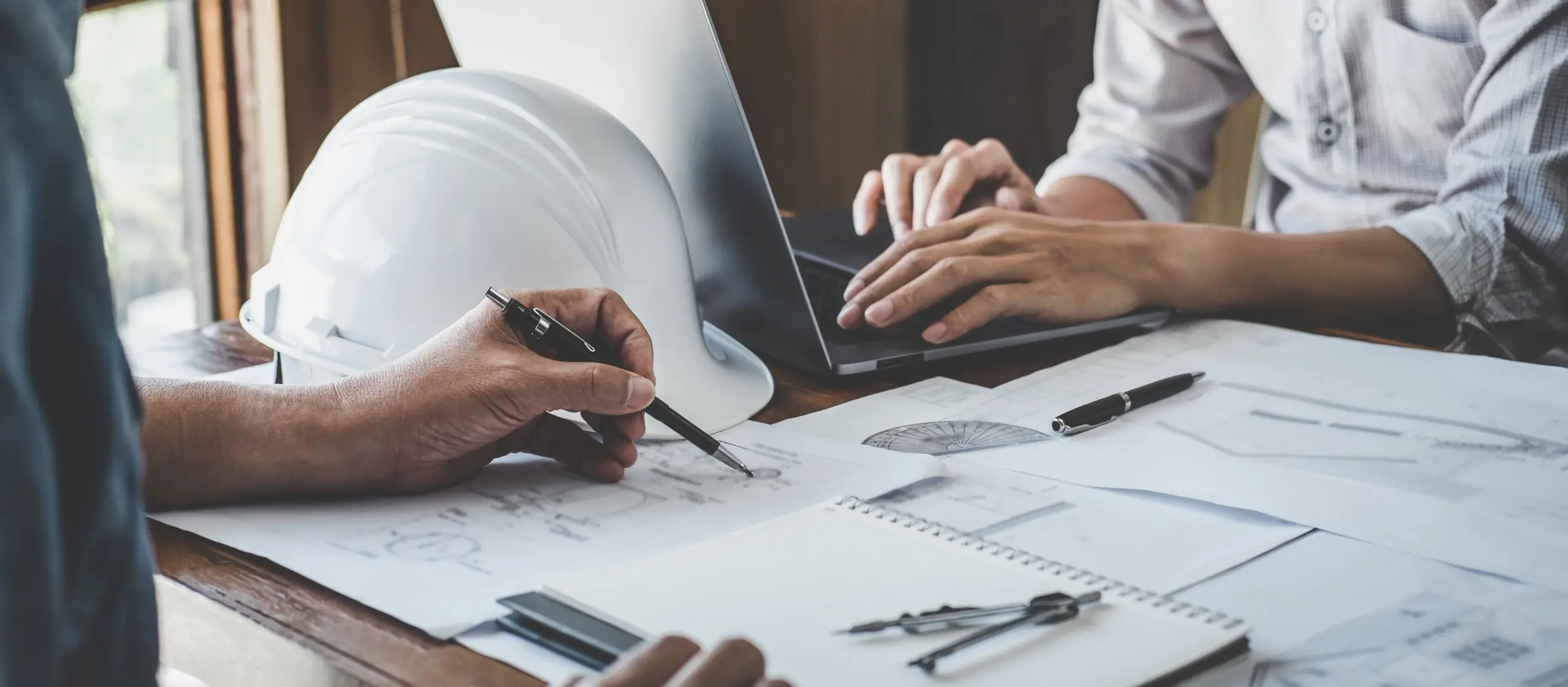 Two people working at a desk with architectural plans, a laptop, a white safety helmet, notebooks, pens, and a compass, suggesting a collaborative engineering or construction project meeting.